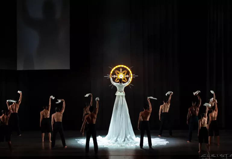 A group of dancers wearing black with their back to the audience face a dancer twice as tall as them wearing a long white dress holding a glowing yellow circle with a star design in the middle.