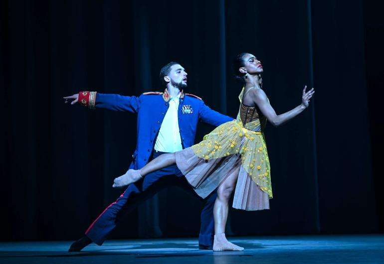 A dancer in a yellow dress strikes an arabesque pose, while a dancer behind holds them by the waist. 