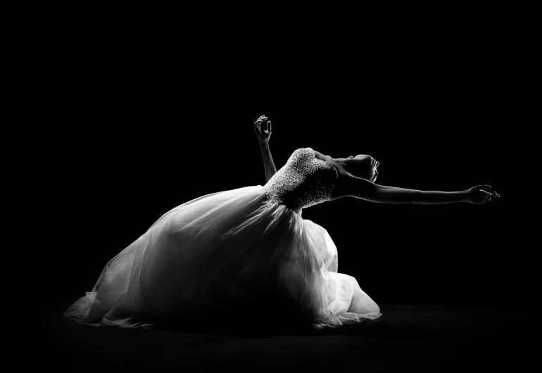 A dancer wearing a long white dress bends their knees and arches their back so their upper body is facing the sky with their arms are out to the side.