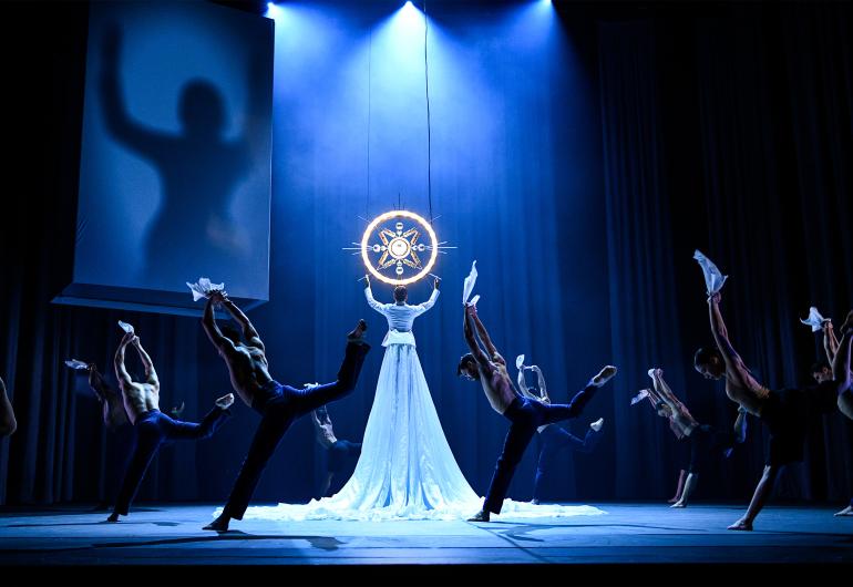 A group of dancers wearing black with their back to the audience face a dancer twice as tall as them wearing a long white dress holding a glowing yellow circle with a star design in the middle. 