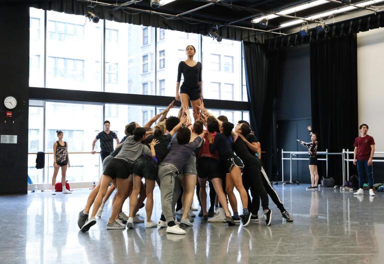 The company in a rehearsal - a tight group reaches up to assist a solo female dancer who is raised above them.
