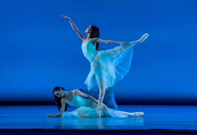 Two female dancers in white, light costumes one a blue lit stage. One in full arabesque and one in pose on the floor.