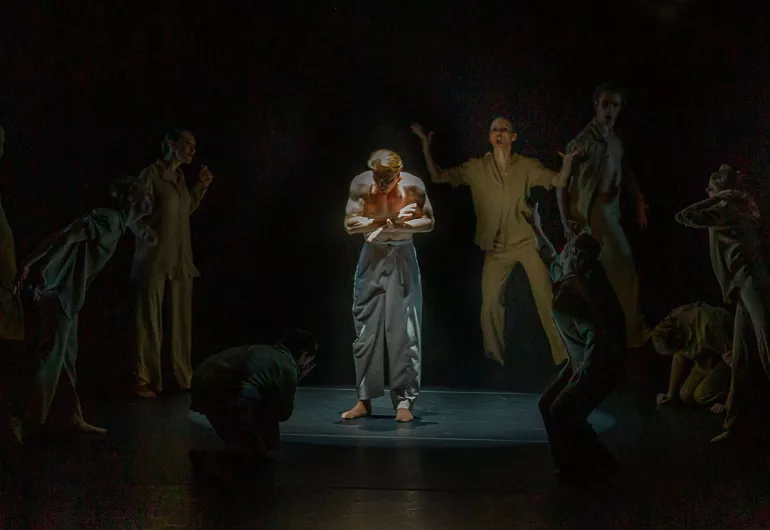 Solo dancer bare chested looking down, suround by people with frantic arms and faces