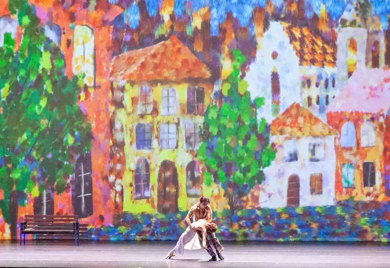 two dancers perform in front of a very color backdrop of a street with houses and trees