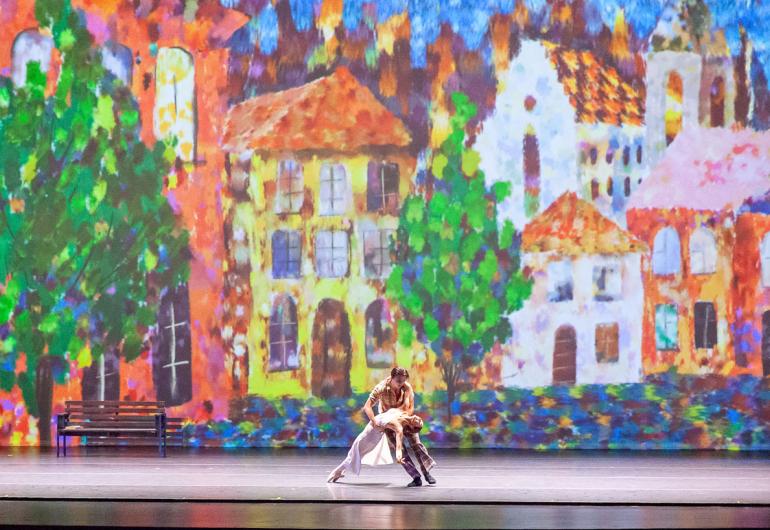 Two dancers on stage in front of a very colorful painting of buildings and trees