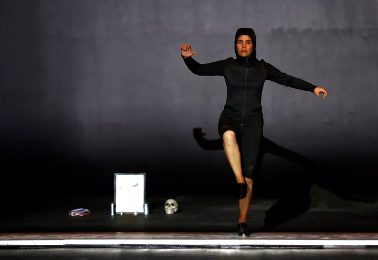 A person wearing all black faces forward with their arms out to the side and right foot off the floor. In the background, there is a red and blue figure, a white rectangle, and a skull.