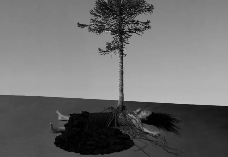 A black and white image depicts a person laying on the floor with their arms overhead. A tree's roots cover the person's body and the tree appears to be coming out of the person's torso.