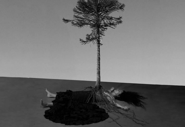 A black and white image depicts a person laying on the floor with their arms overhead. A tree's roots cover the person's body and the tree appears to be coming out of the person's torso.