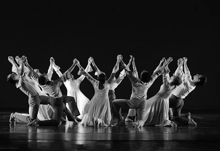 A group of twelve dancers kneel on their right knee with their left foot on the ground in front of them. They are all looking up and holding hands with their arms raised above their heads. This image is black and white.