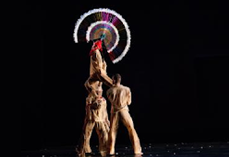 Two dancers hold a third above their shoulders, while a fourth dancers across the stage points a finger at the elevated dancer. The elevated dancer wears a large, circular headpiece, that stands out before the all black background.