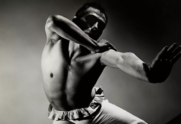 A black and white image of José Limón with their left arm and leg extended and right arm grabbing the left. His head is turned toward his left arm. The background is dark along the edges and lighter in the center.