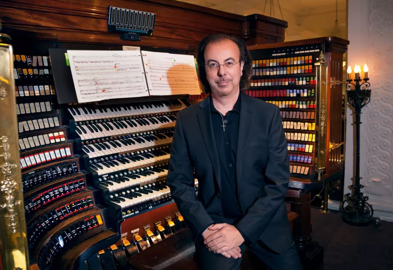 Peter Richard Conte. A black-haired white man wearing glasses and all black sits on a bench with their back to an organ console with their hands crossed on their lap.