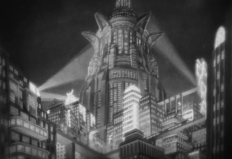 A tall, cylindrical building with many windows is in the middle behind a row of rectangle buildings. Some are lit up and two rays of light shine toward the middle building.