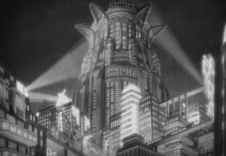 A tall, cylindrical building with many windows is in the middle behind a row of rectangle buildings. Some are lit up and two rays of light shine toward the middle building. 