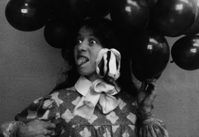 A young person looking to their right with wide eyes sticking their tongue out. They are holding balloons and have bows in their hair.