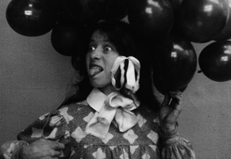A young person looking to their right with wide eyes sticking their tongue out. They are holding balloons and have bows in their hair. 