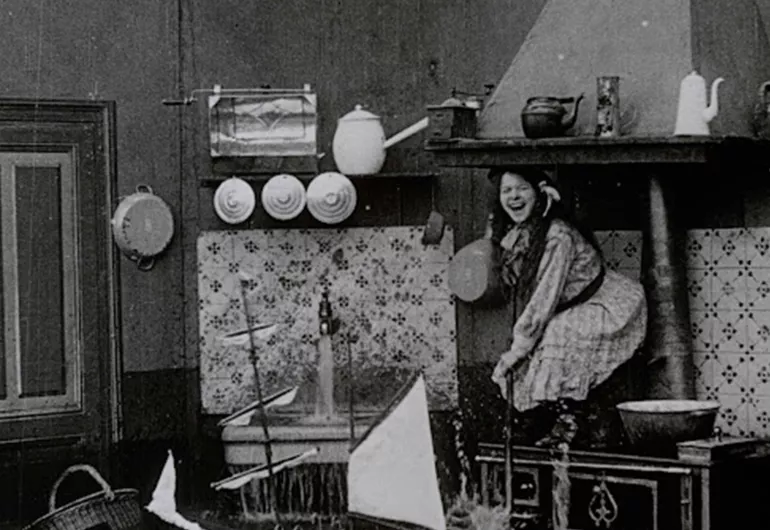 A person is crouching on a raised surface laughing. There is a running sink to their right and the floor appears to be flooded. Behind the person is a white and black patterned wall.