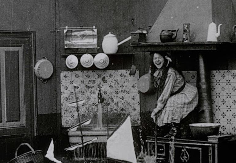 A person is crouching on a raised surface laughing. There is a running sink to their right and the floor appears to be flooded. Behind the person is a white and black patterned wall. 