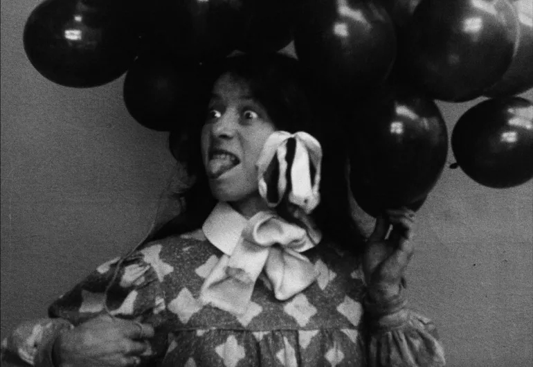 A young person looking to their right with wide eyes sticking their tongue out. They are holding balloons and have bows in their hair. 