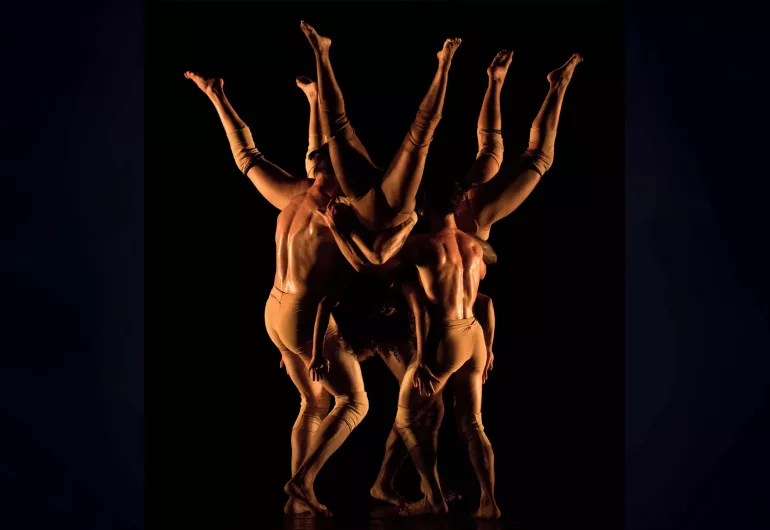 On a dimly lit stage, three dancers are held upside down in a circle while three other dancers hold them.