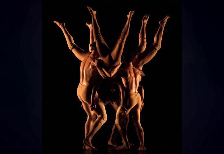 On a dimly lit stage, three dancers are held upside down in a circle while three other dancers hold them. 