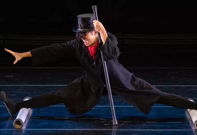 A person's legs are on two cylinders completely spread apart and nearly doing the splits. They are wearing a black hat and holding a cane.