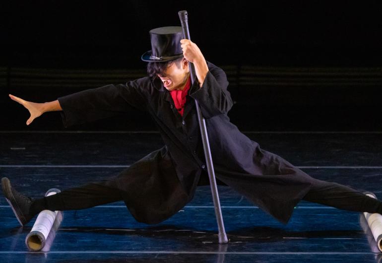 A person's legs are on two cylinders completely spread apart and nearly doing the splits. They are wearing a black hat and holding a cane. 