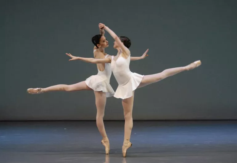 Two dancers stand side by side holding each other's left hand above their head. One leg is extended behind them. Their dresses are short and white. The background is grey and blue.