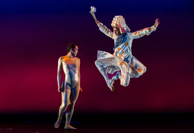 Image of a stage shows two dancers, one standing and the other leaping in the air with their hands above their head.