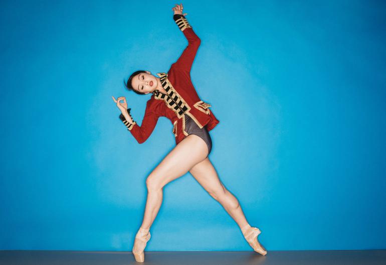 Caili Quan on pointe; wearing a drum major jacket; legs in a wide step forward, body twisted to side and tilted with angled raised arms