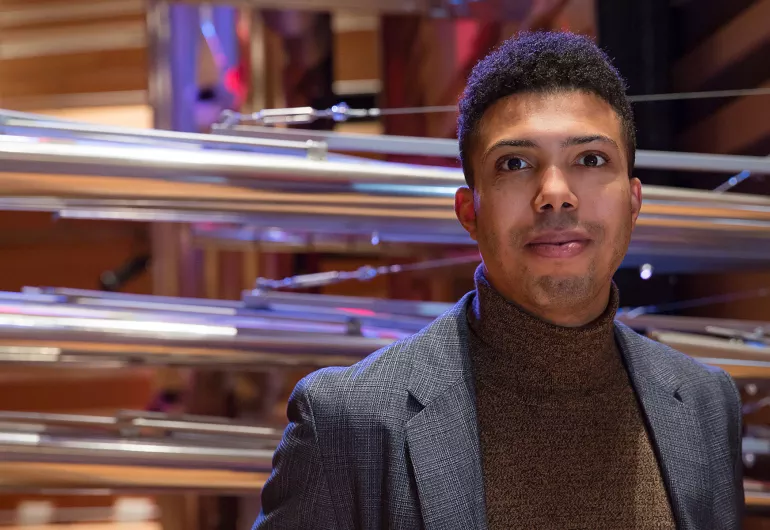 A person with dark hair wearing a brown turtleneck under a dark gray suit coat stands toward the camera with large organ pipes behind them.