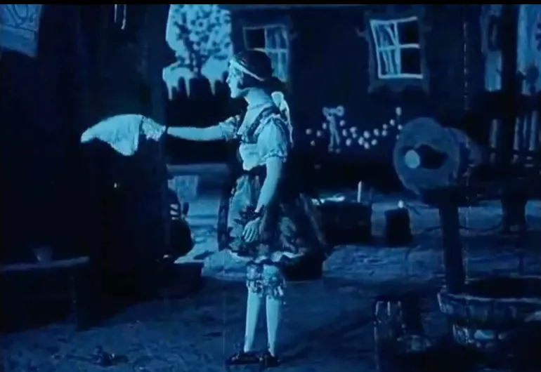 A black and white image tinted blue shows a person wearing a dress and a hairband, standing outside with their body facing to the left and arm extended outward.