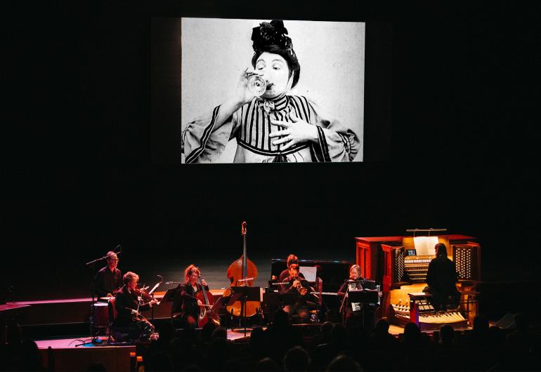 Musicians in the foreground playing in front of a movie screen showing a black and white image of a woman drinking from a wine glass.