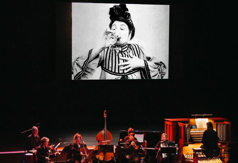An ensemble of 7 musicians dressed in black perform onstage with a projection of a film above them. The screen shows a black and white film still of a person wearing a striped top, drinking a glass of wine with one hand held to their chest and their eyebrows raised.