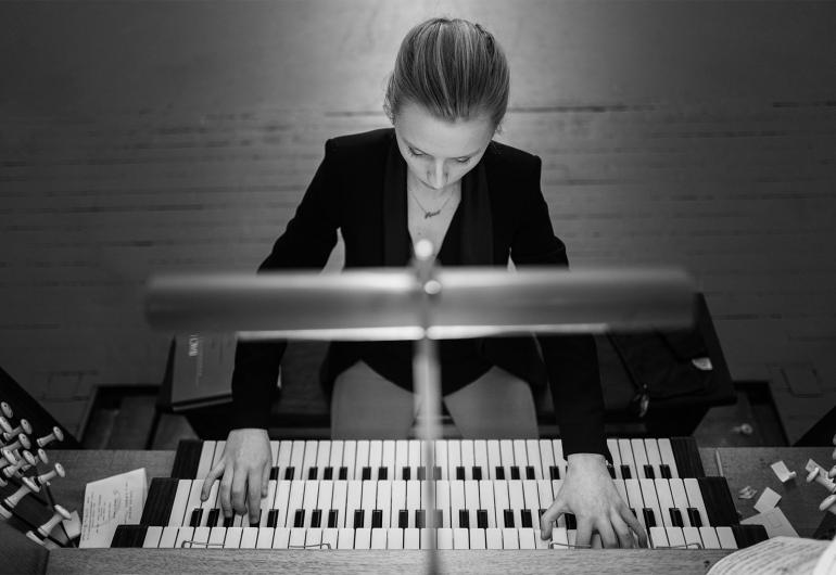 A black and white image shot from above shows a person wearing a blazer looking down and playing a pipe organ. 