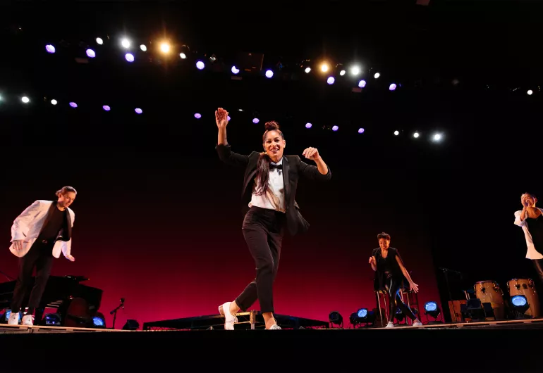 Four dancers are onstage dressed in tap shoes with black pants and a mix of black and white tops. One dancer is slightly downstage, while the other Three dancers form a semicircle around them.