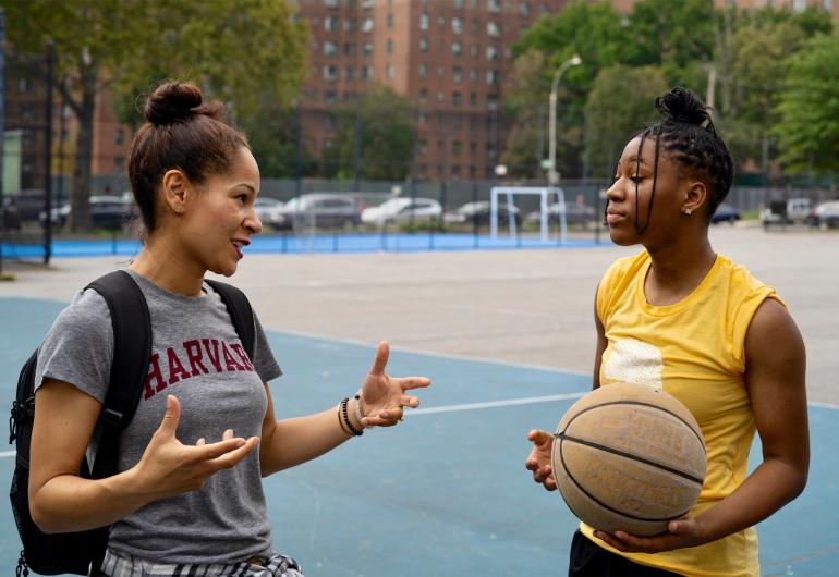 Casel in casual conversation with a young woman while standing on an outdoor basketball court.