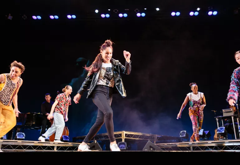 Five dancers and one musician appear in-motion onstage in casual clothing. Three dancers are slightly downstage, while the other three dancers and one musician are somewhat behind them on a raised platform.