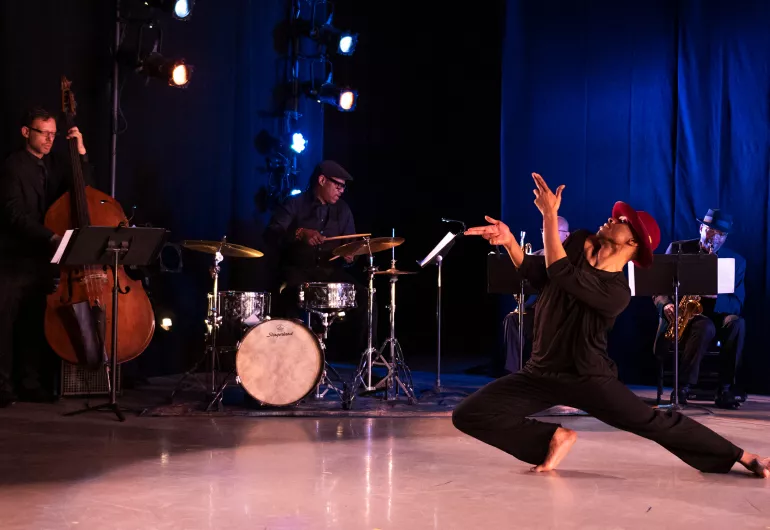 1 dancer dressed in black with red headwear is positioned onstage in front of a bassist, drummer, and saxophonist performing behind them. They face to the left with their arms pointed up, and one leg bent and the other extended outward.