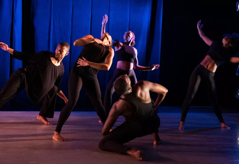 A group of 5 dancers wearing black clothing appear onstage with cobalt blue lighting. 3 dancers stand facing the camera with their arms extended upward and parallel to the floor while 1 dancer kneels, facing the curtain.