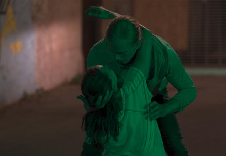 A couple embrace/dance closely in an urban setting. They are bathed in a green light.