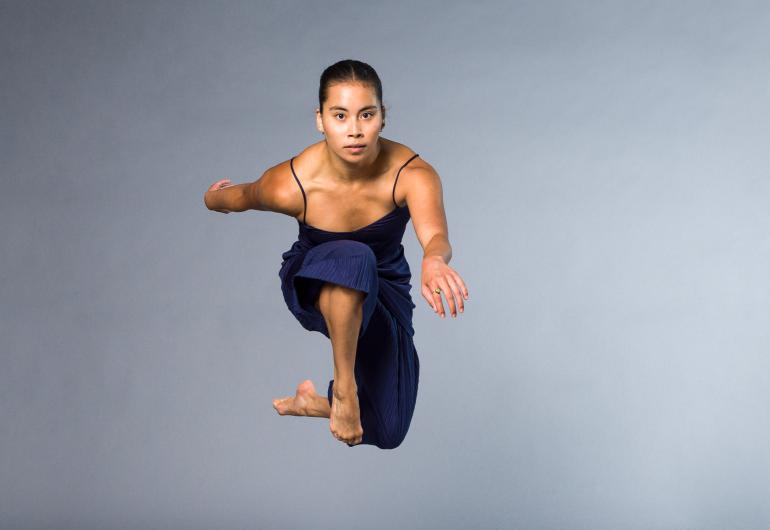A dancer dressed in blue appears in front of a gray backdrop, jumping in the air and facing directly at the camera.