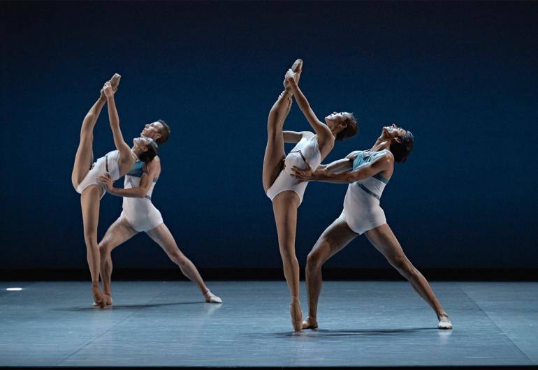 Two pairs in white leotards, female dancers on pointe while holding the other leg straight up toward their head