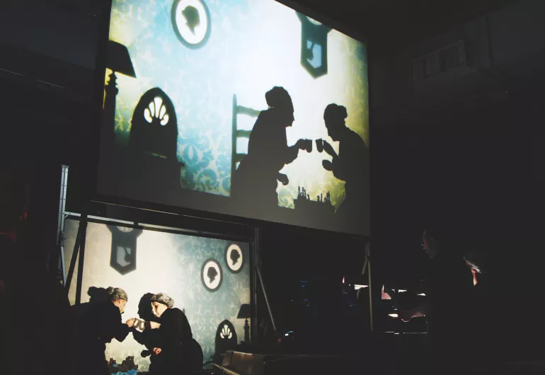 Actors portray 2 elderly women having coffee in the show film projected above the scene