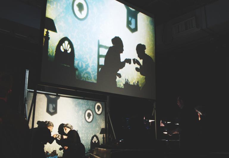 Actors portray 2 elderly women having coffee in the show film projected above the scene