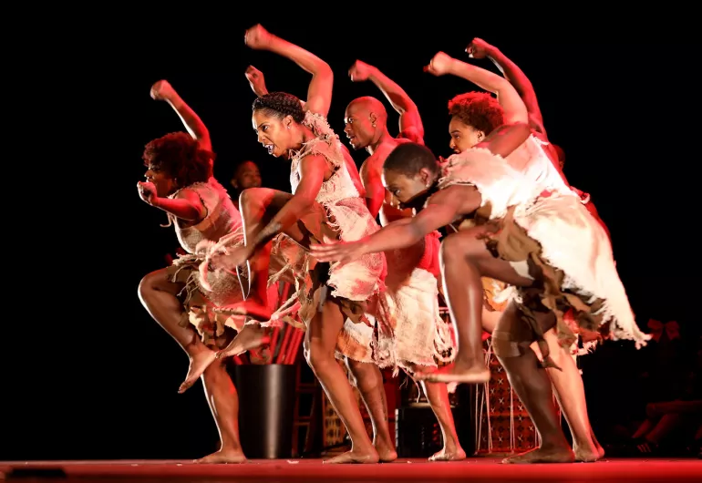 A group of dancers stand closely together onstage dressed in white clothing with their right arms and left legs raised, gazing downward toward the left and accompanied by a soft glow of red lighting.