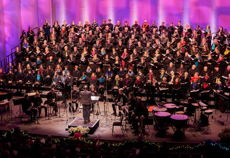 A choir wearing black with various colored scarves performs onstage with patterned pink and purple lighting projected behind them. A conductor stands downstage with their back toward the camera and musicians surrounding them.