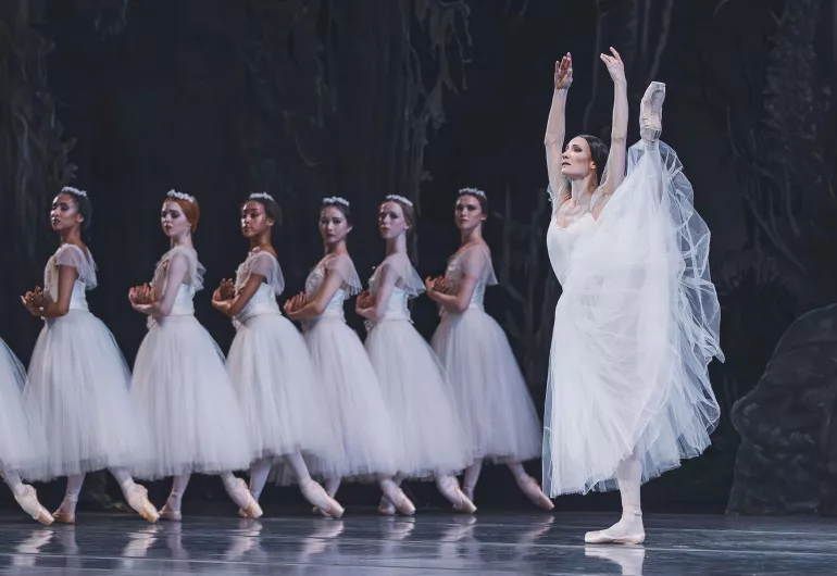 A ballerina extends her leg to the side above her head. She stands beside a line of identically posed dancers in white.