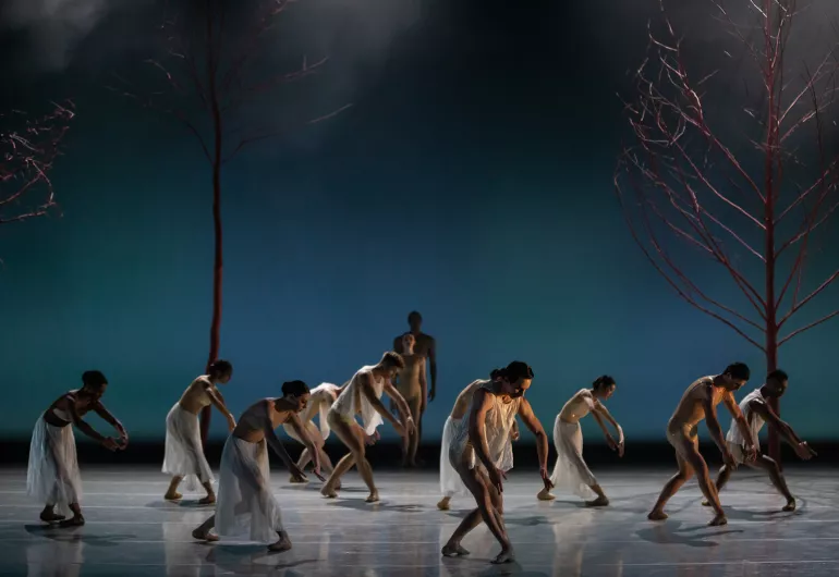 BalletMet dancers in white costumes gracefully bent over onstage by tree scenery.