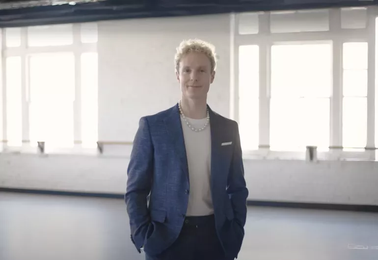 Remi Wörtmeyer, a man with curly blonde hair, smiles in a blue suit and pearl necklace.